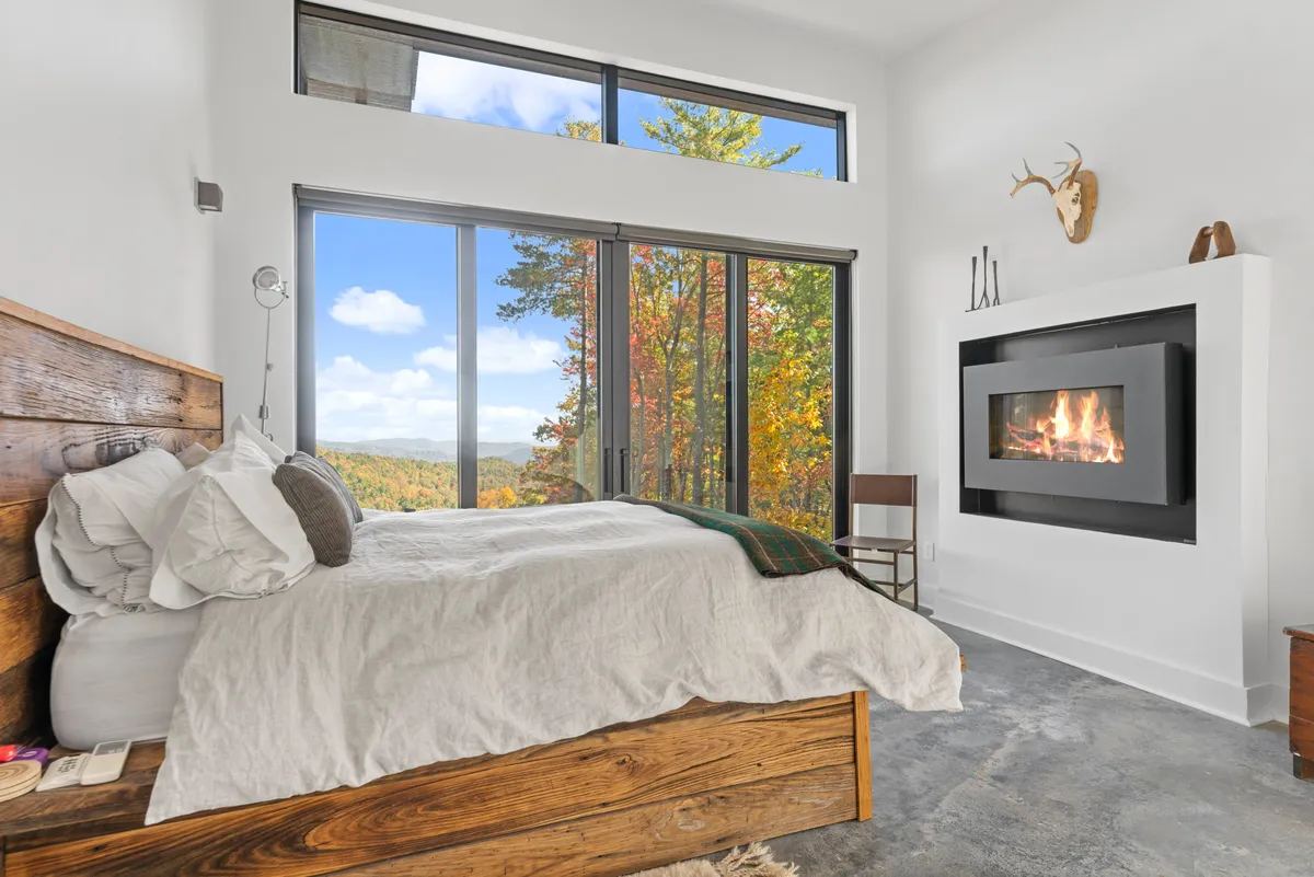 This image captures a cozy bedroom with a large window framing vivid autumn foliage, a modern fireplace, and layered bedding. Natural wooden accents in the furniture enhance the sense of warmth and connection to nature. The outside world is brought in through the broad view of the changing leaves, inviting guests to enjoy the seasonal beauty from the comfort of indoors.