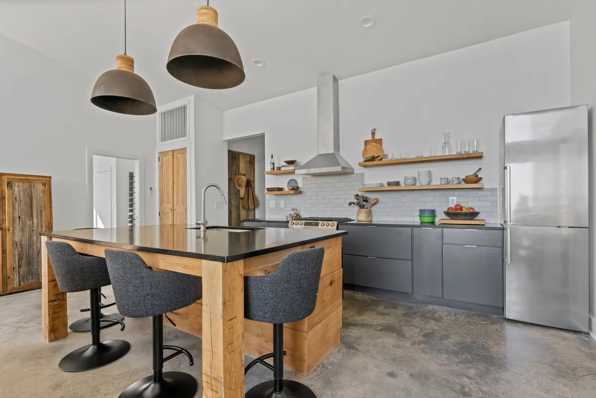 A contemporary kitchen featuring a large kitchen island as the centerpiece, illuminated by stylish hanging pendant lights. Open shelving displays ample kitchenware and cookware, while the overall space is bathed in natural light. The setup encourages both socializing and culinary creativity.