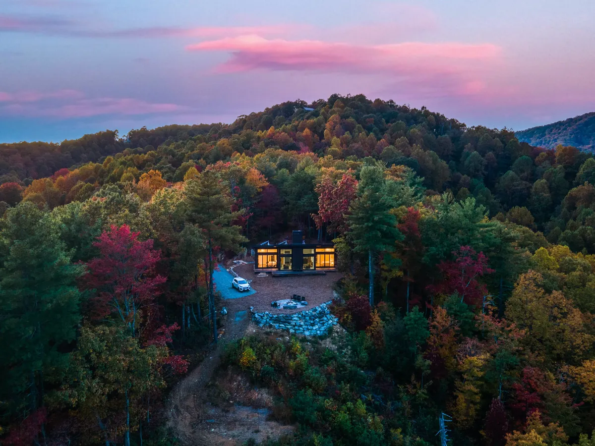 A modern house nestled among colorful autumn foliage, set against the dramatic backdrop of the Blue Ridge Mountains. The driveway leads up to the home, and the tranquility of the setting suggests easy access to nature trails and outdoor adventures.