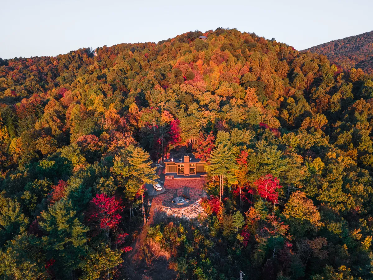 The house sits at the edge of a wooded area, with a cozy fire pit surrounded by chairs and the forest enveloping the space. The setting is perfect for outdoor gatherings, quiet evenings, or starting a hike into the surrounding trails.
