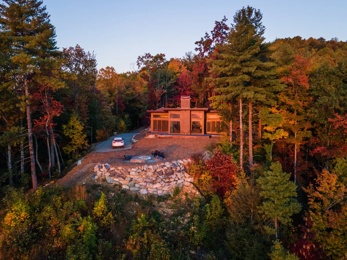 This exterior image captures a modern home nestled among trees with a prominent fire pit in the foreground. The setting is tranquil, ideal for outdoor gatherings, with ample space for relaxing or stargazing around the campfire. The twilight ambiance suggests a cozy, romantic evening under the stars.