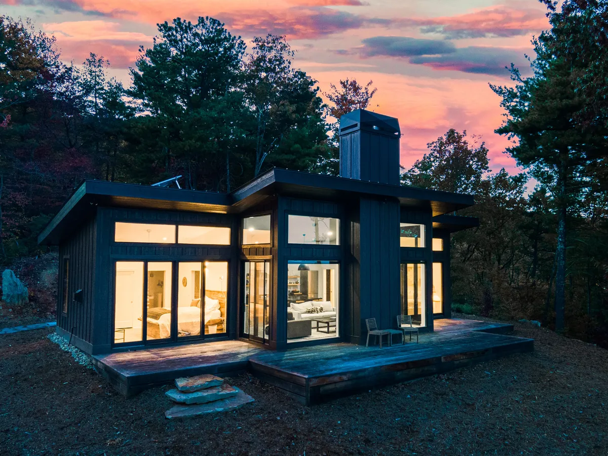 The image showcases the modern, architecturally striking exterior of a luxury tiny house surrounded by lush trees, featuring expansive glass windows and a spacious wooden deck. The setting is serene and private, with the deck perfectly positioned for guests to relax and soak in breathtaking long-range mountain views, ideal for sunset watching or morning coffee. The overall mood is tranquil, cozy, and invites outdoor connection with nature.