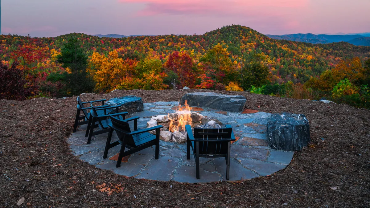 An inviting outdoor campfire area surrounded by comfortable chairs, set against a backdrop of autumn foliage and expansive mountain views. The warm firelight glows as dusk falls, encouraging gathering, storytelling, and stargazing.