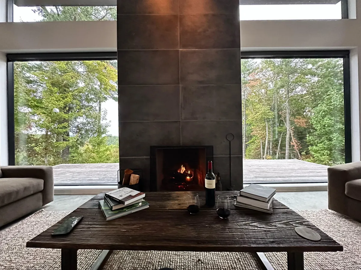 An alternate angle of the great room, focusing on the interplay between the fireplace, large windows, and comfortable sofas. The wooden table and soft lighting evoke a welcoming setting ideal for both social gatherings and quiet reflection, all with the outdoors visible beyond.