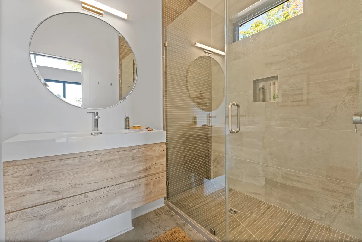 A modern bathroom with a circular mirror above a wooden vanity, a spacious glass shower enclosure, and clean lines. The serene palette and elegant finishes evoke a spa-like atmosphere.