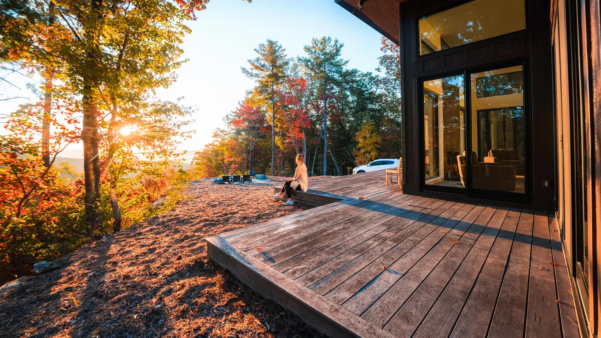 A calm, sunset-lit deck scene with a person sitting in comfortable outdoor seating, surrounded by trees and open sky. The deck extends out into the natural surroundings, offering an immersive connection to the landscape and a sense of tranquility.