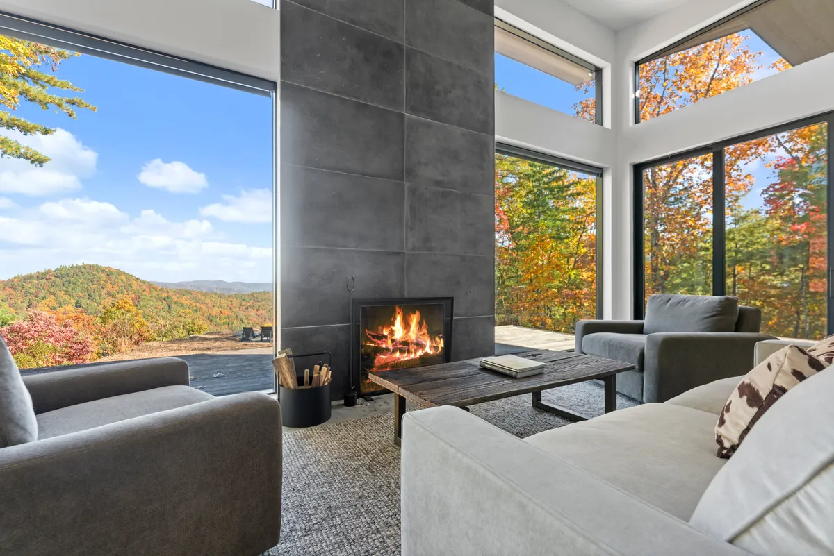 This image showcases the great room’s interior with a wood-burning fireplace, generous coffee table, plush sofas, and dramatic floor-to-ceiling windows revealing sweeping mountain views. Sunlight fills the space, creating a warm, inviting atmosphere that encourages relaxation and conversation.