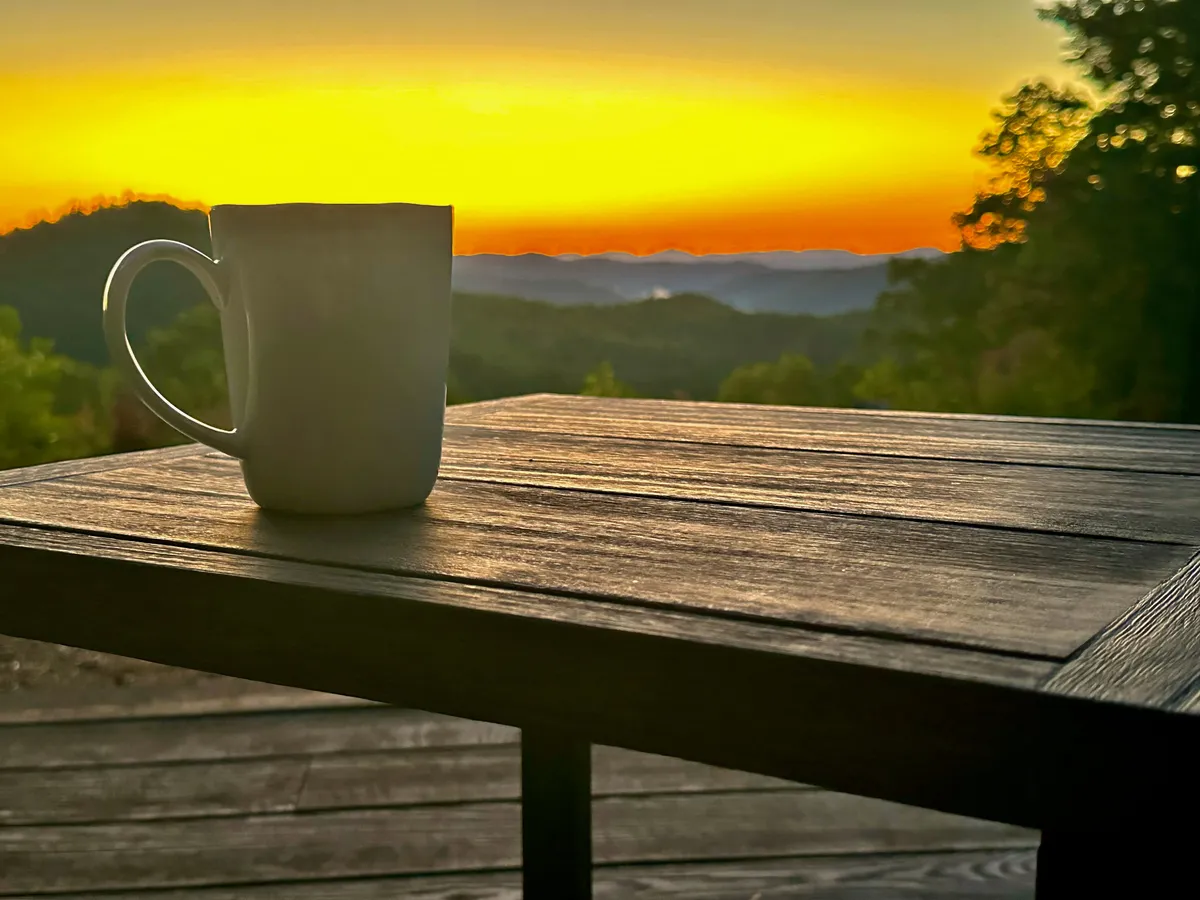 A steaming coffee mug sits on a wooden deck railing, overlooking a sweeping mountain range bathed in sunrise or sunset hues. The scene is tranquil, inviting contemplation and connection with nature.