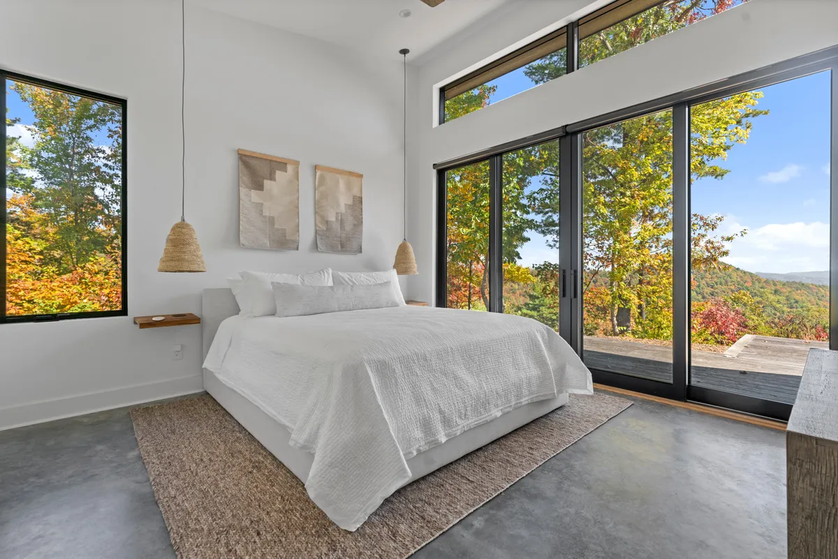A serene bedroom featuring a large, plush bed with crisp white linens, floor-to-ceiling windows offering direct access to a private deck, hanging light fixtures, and textile wall art. Natural light fills the space, with views of the outdoors visible beyond the windows.