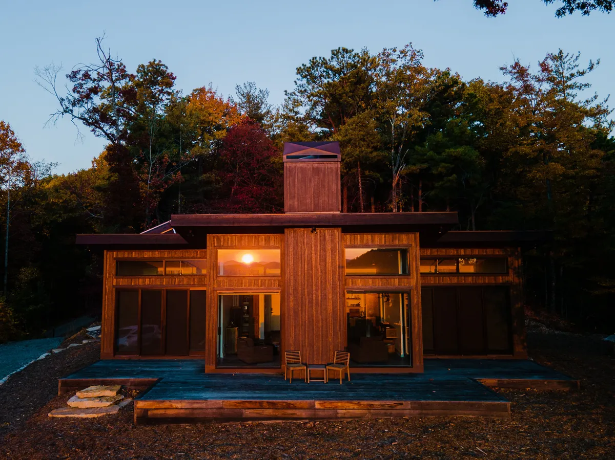 The photo features a modern wooden house with expansive windows, surrounded by trees and bathed in a golden sunset glow. The inviting deck and serene setting highlight the blending of indoor and outdoor relaxation, with spaces perfect for unwinding after a day of exploring nearby towns.