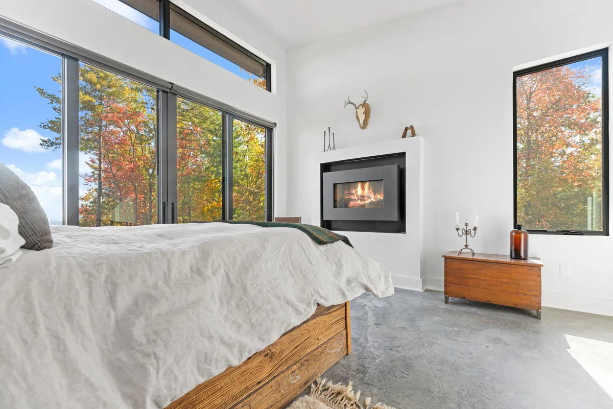 A calm, inviting bedroom with a cozy bed, a sleek gas fireplace, and windows offering mountain and nature views. The room’s neutral palette creates an atmosphere ideal for relaxation after a day of adventure.